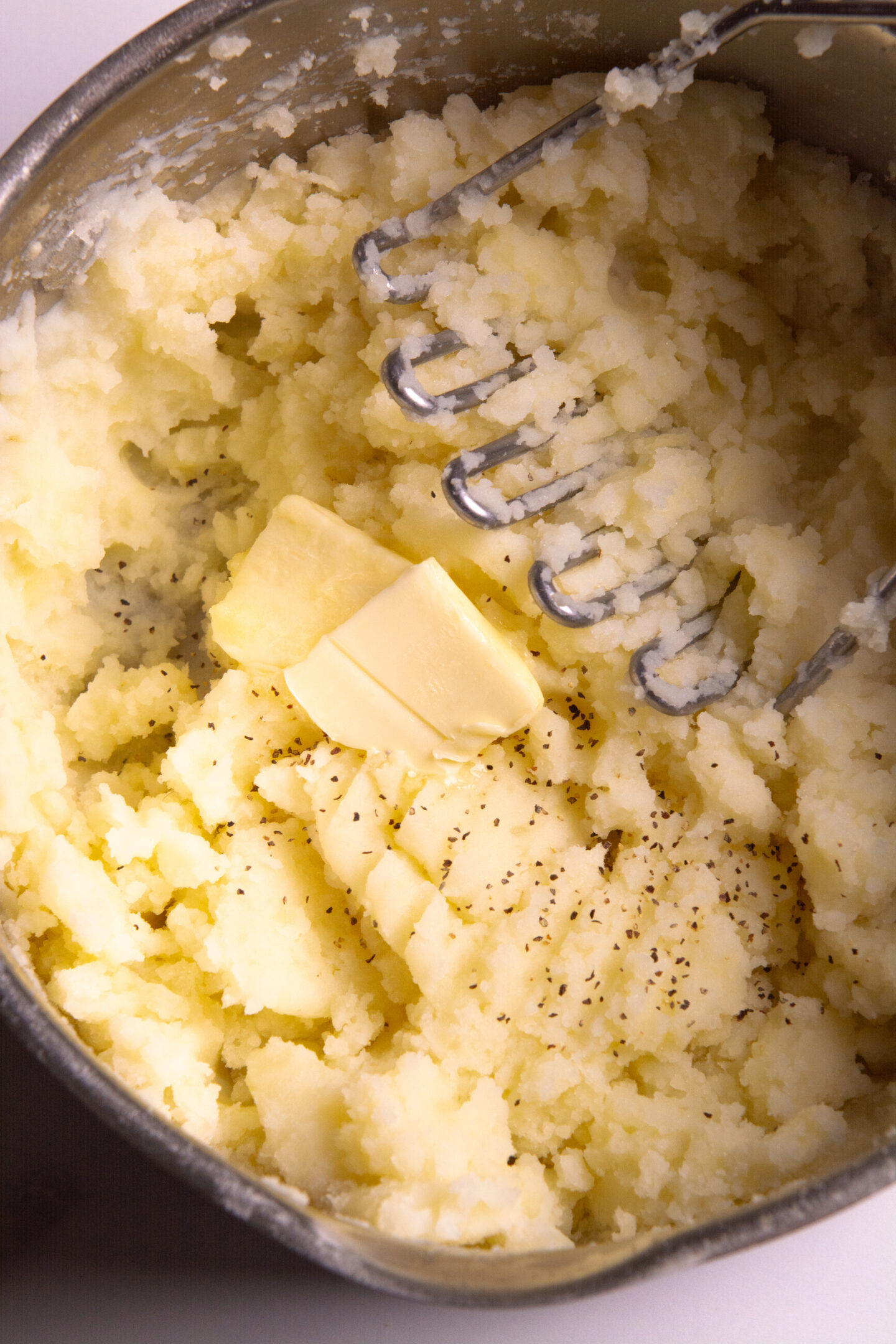 A pot of potatoes being mashed with a potato masher, topped with two tablespoons of butter and salt and pepper.