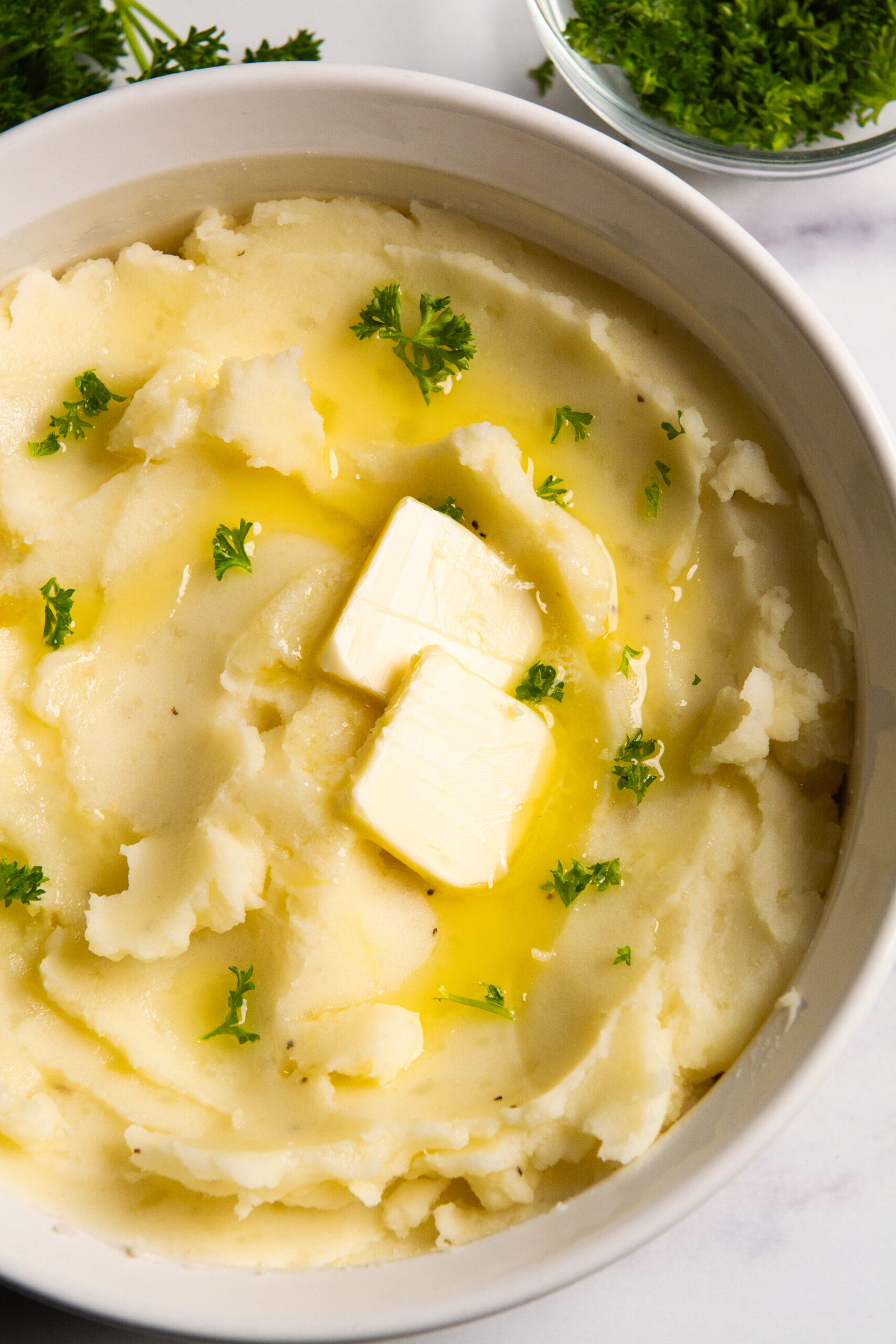 A bowl of mashed potatoes with two tablespoons of butter, drizzled with more melted butter and sprinkled with parsley.