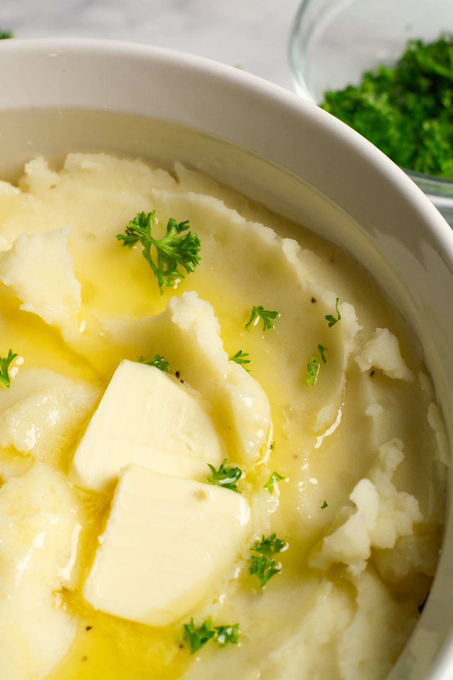 A close up of a bowl of mashed potatoes with two tablespoons of butter, drizzled with more melted butter and sprinkled with parsley.