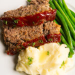 A plate with meatloaf and a sweet glaze, with green beans and mashed potatoes