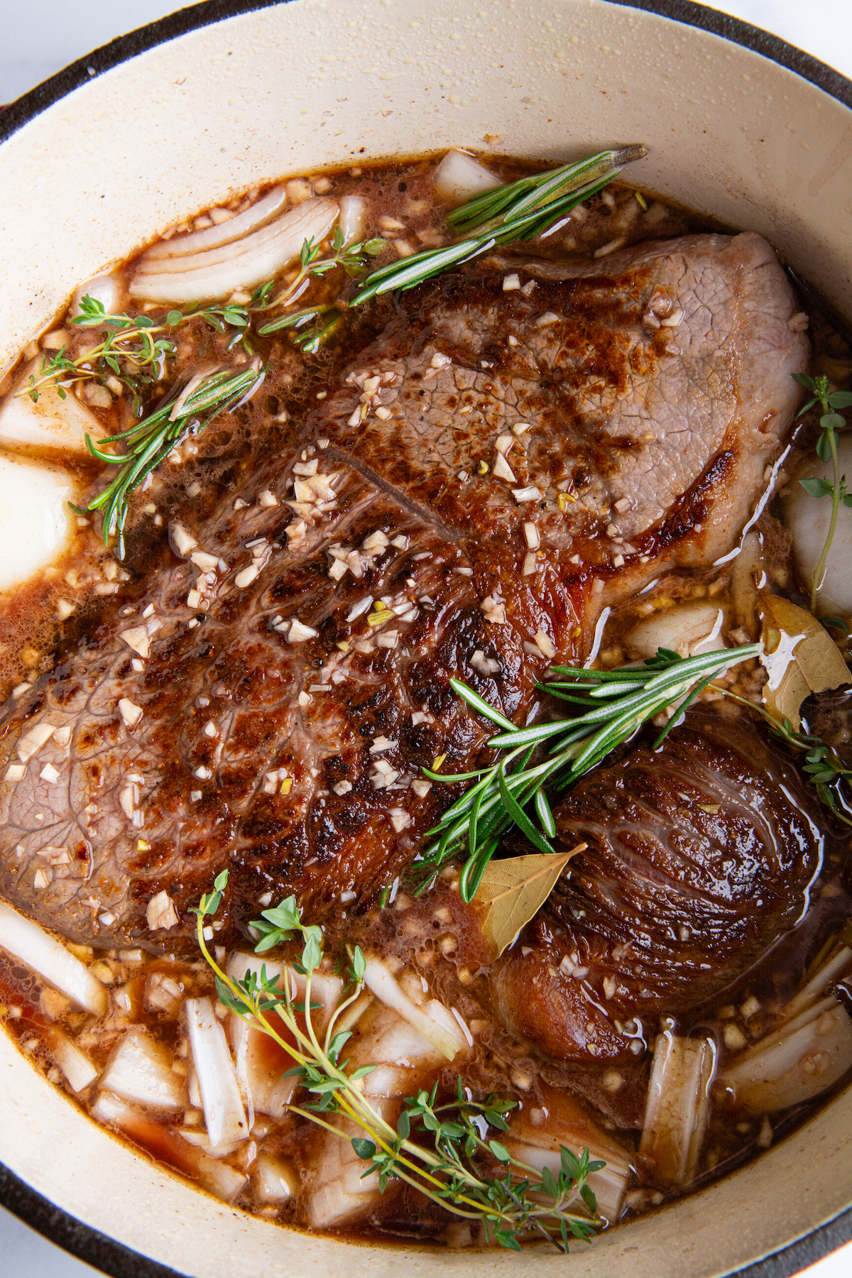 A large Dutch oven with seared rump roast, chunks of white onion, sprigs of thyme and rosemary, covered with beef stock and red wine.