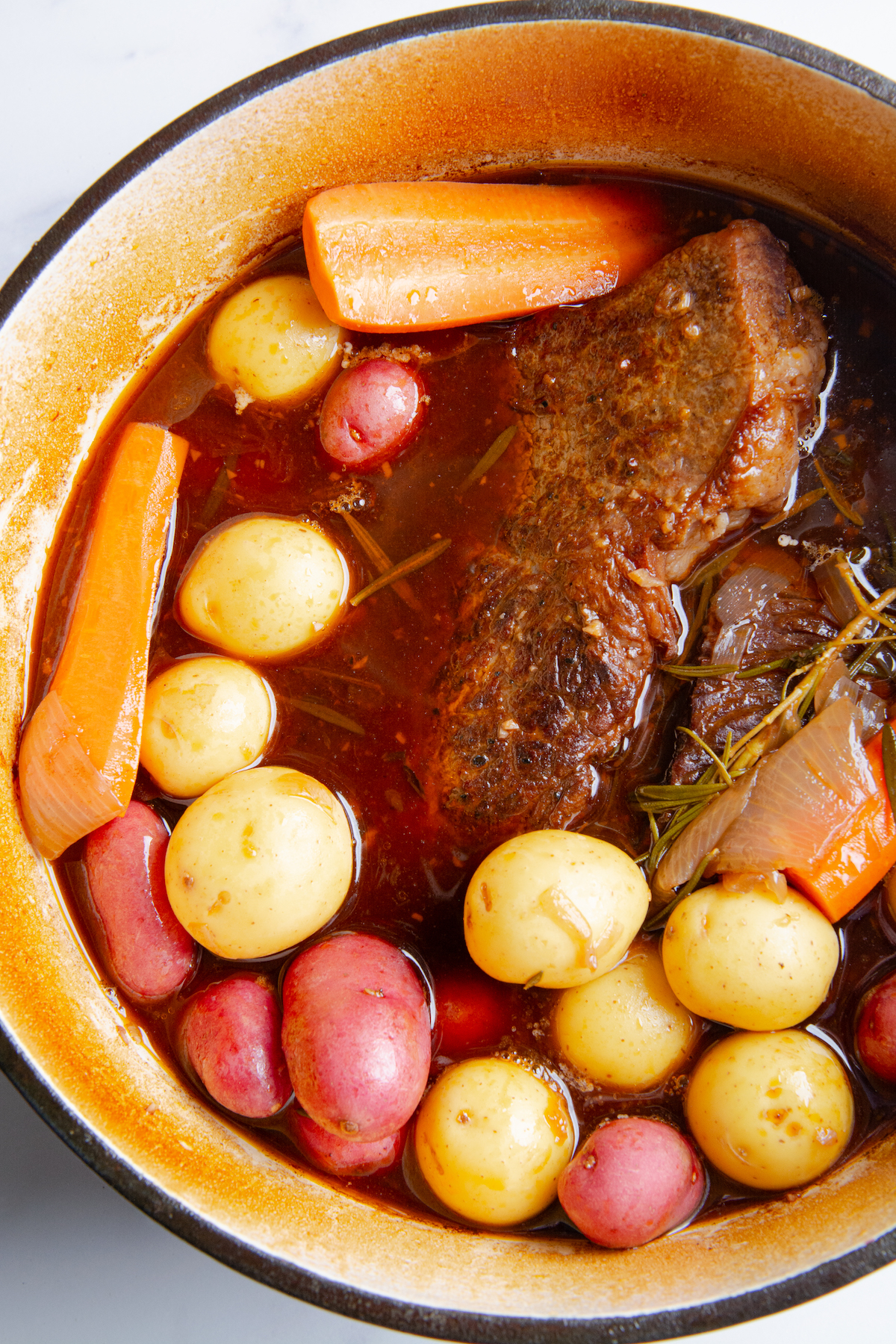 A large Dutch oven with a partially cooked rump roast in beef stock and red wine, with added baby potatoes and carrots.