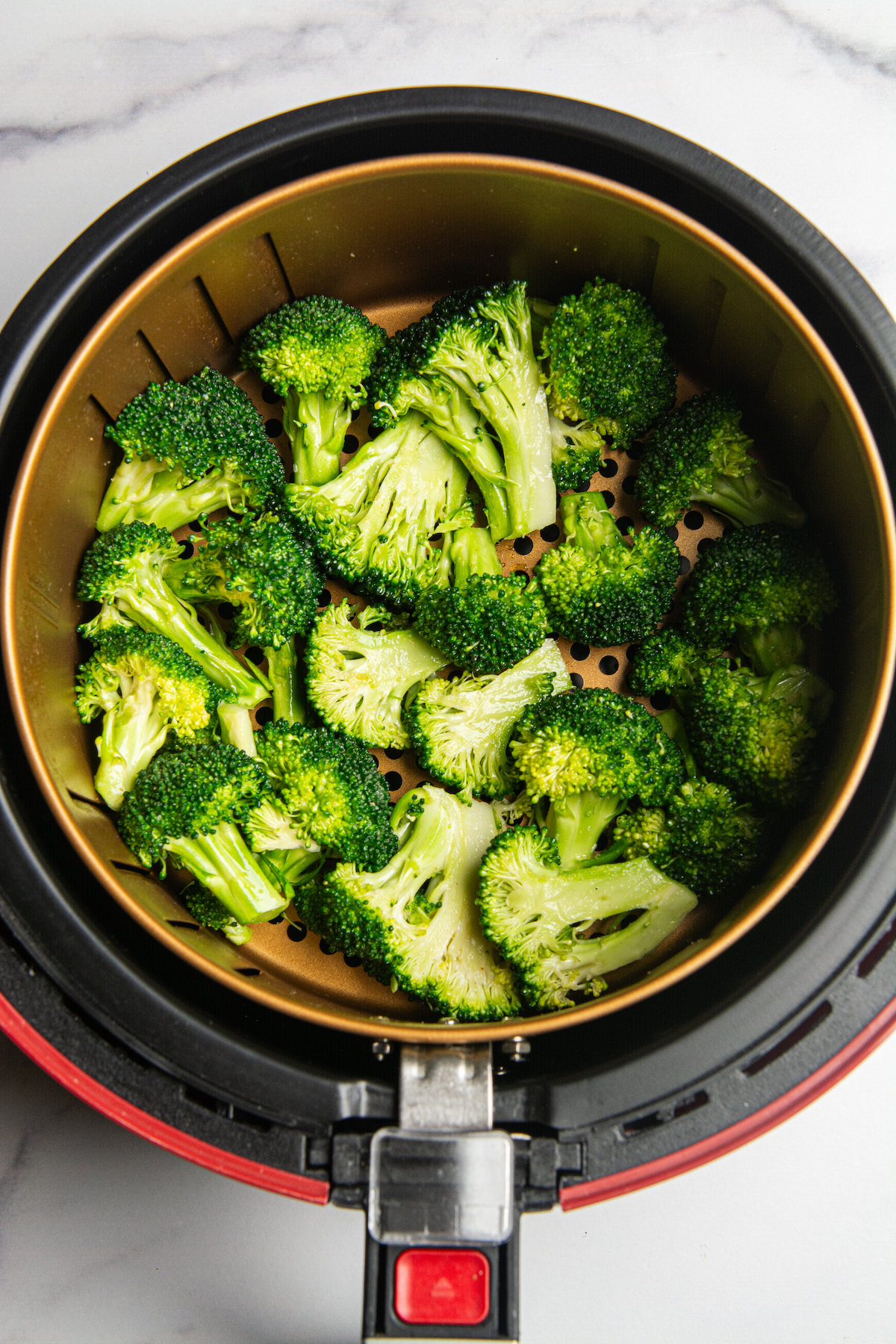 An air fryer with a basket of raw and seasoned broccoli florets.