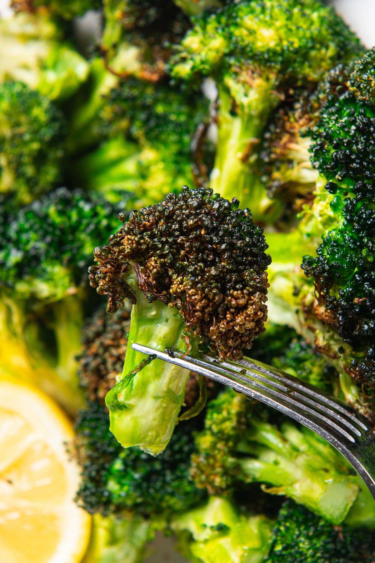 A fork holding a roasted broccoli floret with roasted broccoli and a lemon wedge in the background.
