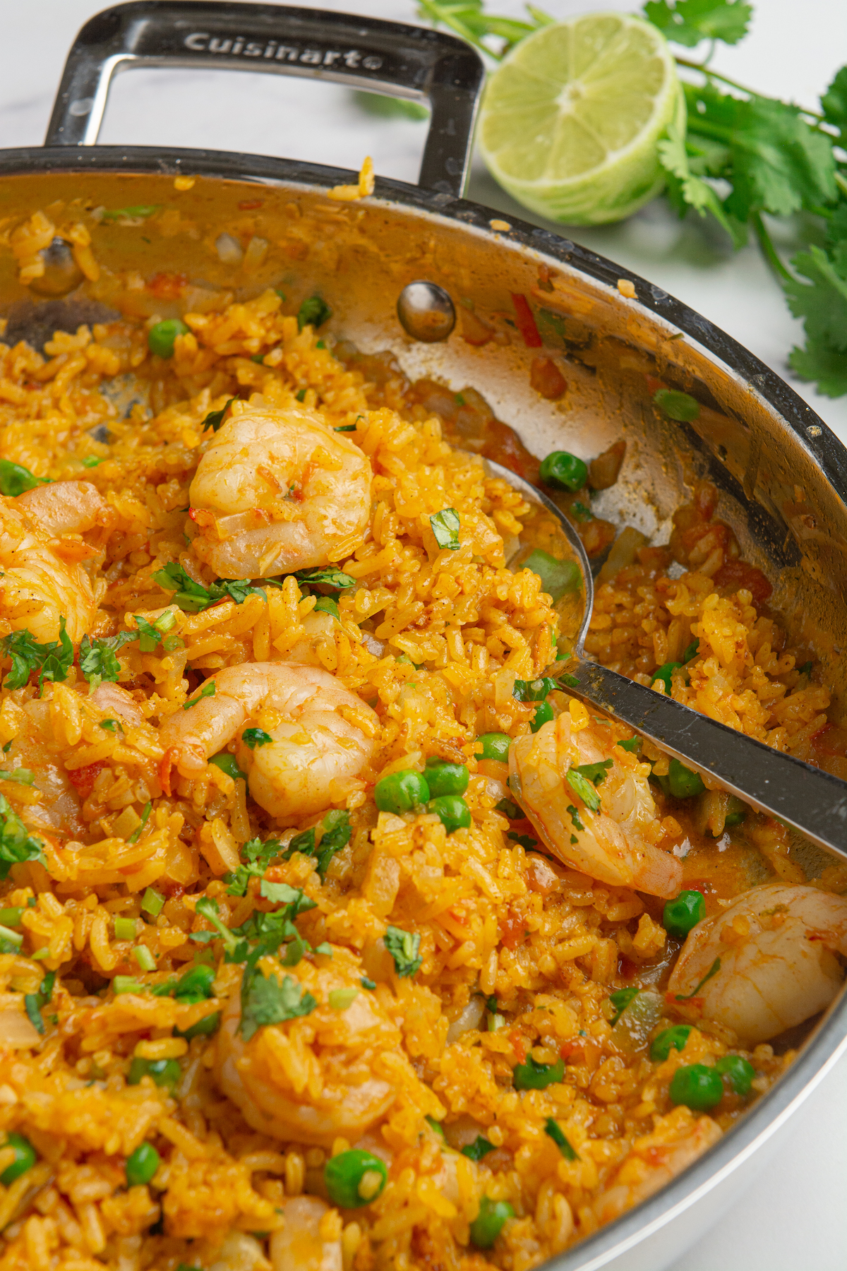 A large stainless steel skillet with cooked arroz con camarones or shrimp and rice with cilantro and sweet peas. Half a lime and a bunch of cilantro in the background.