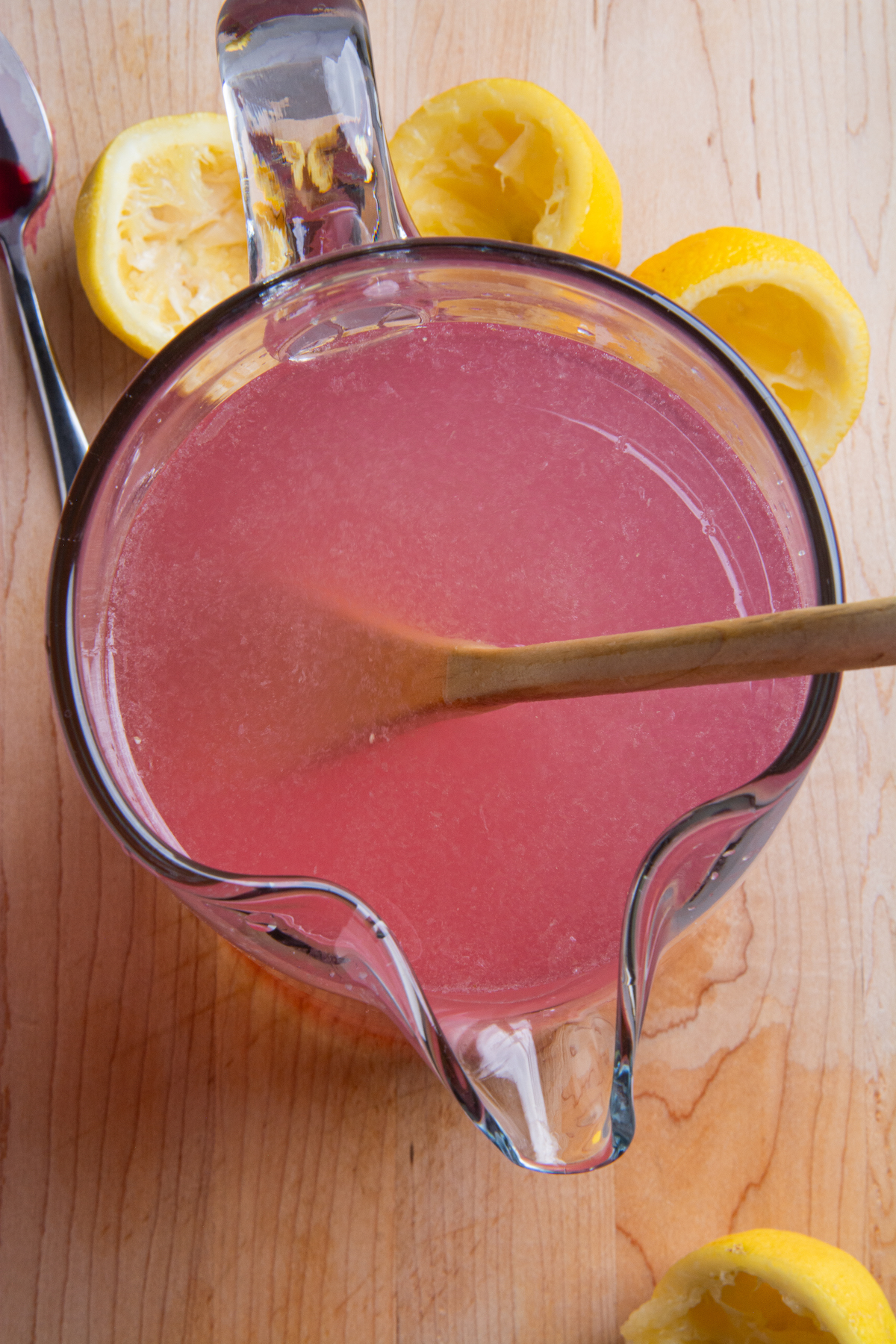 Above a pitcher of pink lemonade with a wooden spoon inside to stir it and squeezed lemons at the bottom around it.