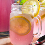 A glass of pink lemonade with lemon slices with a pitcher of pink lemonade in the background.