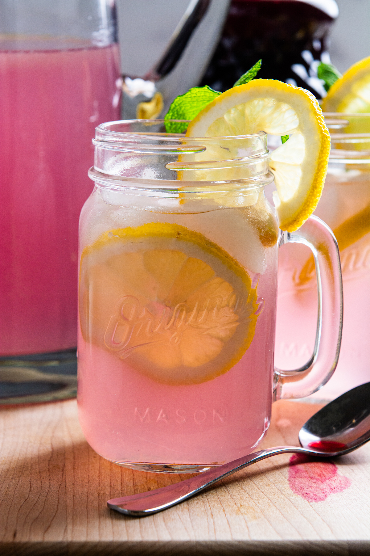 A glass of pink lemonade with lemon slices with a pitcher of pink lemonade in the background.