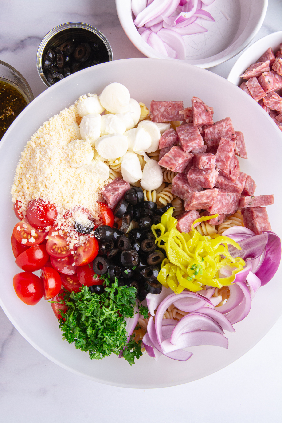 A bowl of ingredients for pasta salad. Cooked rotini pasta, sliced cherry tomatoes, chopped parsley, sliced red onion, sliced pepperoncini, sliced black olives, chopped dry salami, mozzarella balls, grated Parmesan cheese.