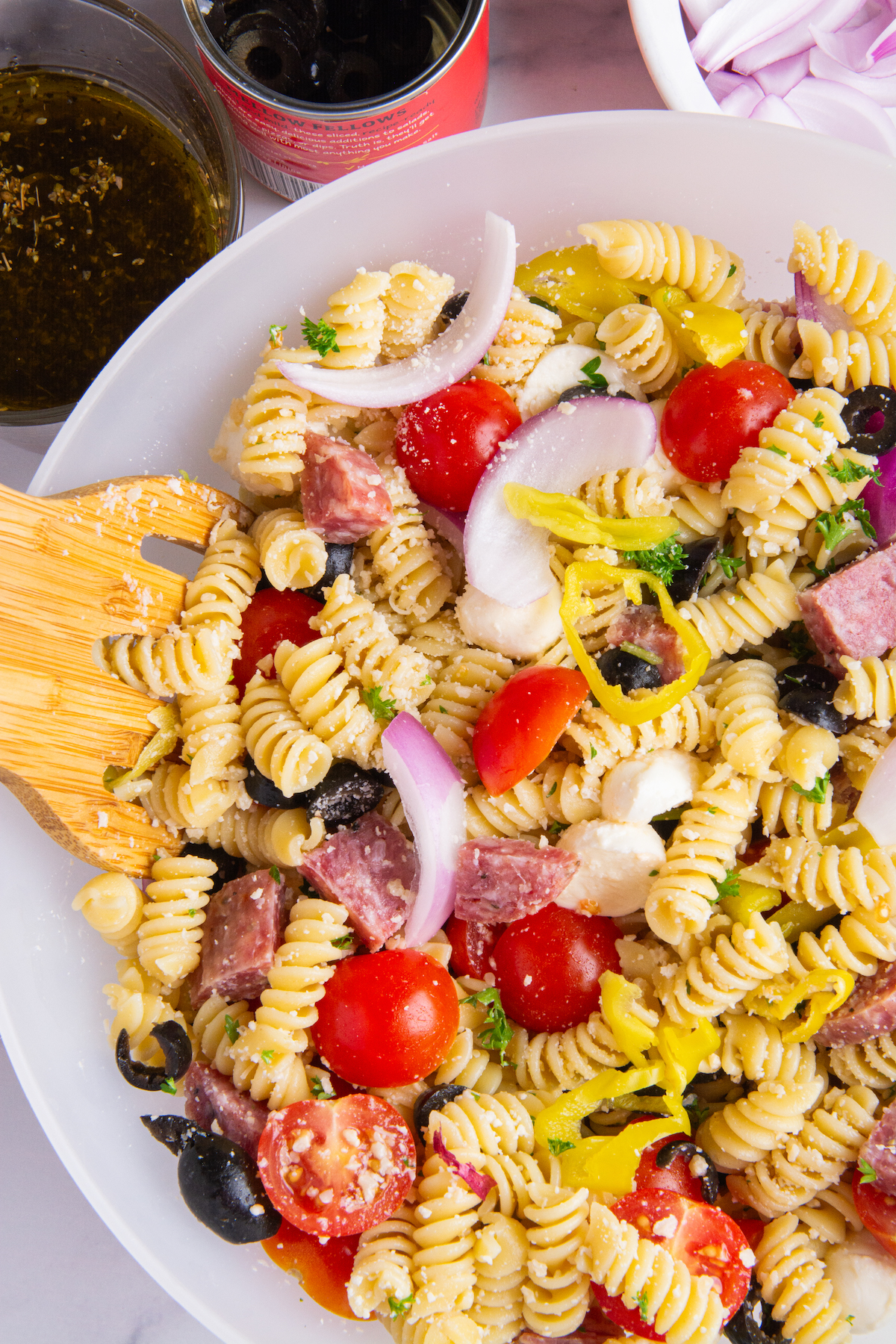 A bowl of Italian pasta salad without dressing. Cooked rotini pasta, sliced cherry tomatoes, chopped parsley, sliced red onion, sliced pepperoncini, sliced black olives, chopped dry salami, mozzarella balls, grated Parmesan cheese.
