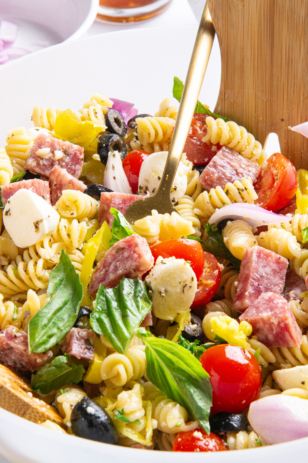A bowl of Italian pasta salad with a gold fork. Ingredients include cooked rotini pasta, sliced cherry tomatoes, chopped parsley, sliced red onion, sliced pepperoncini, sliced black olives, chopped dry salami, mozzarella balls, grated Parmesan cheese.
