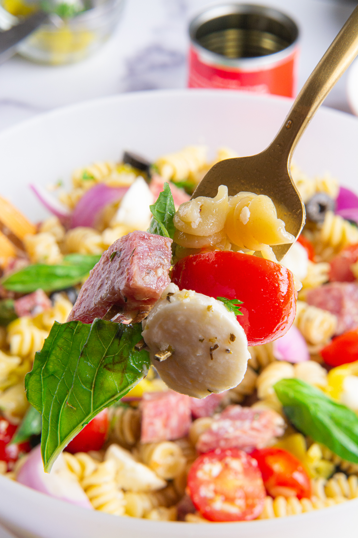 A fork holding cooked rotini pasta, a cherry tomato, dry salami, mozzarella cheese and a basil leaf. The Italian pasta salad can be seen in the background.