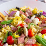 An Italian salad bowl with rotini pasta, cherry tomatoes, mozzarella, black olives, salami, pepperoncinis, and basil leaves.