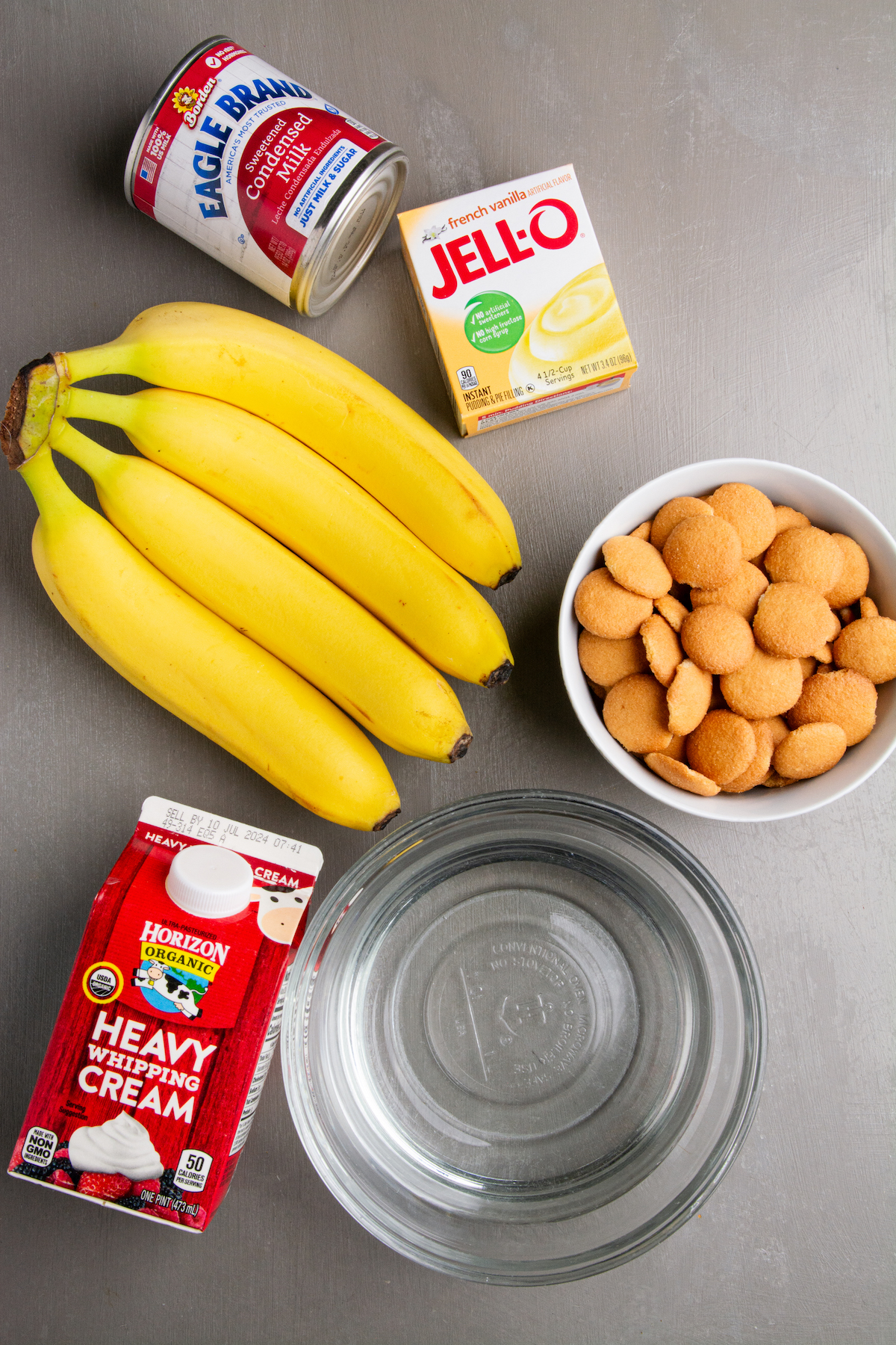 Ingredients for Magnolia Bakery's Banana Pudding. A bunch of bananas, a can of condensed milk, a box of instant French vanilla pudding, a bowl full of Nilla wafers, a bowl of cold water, and a small carton of heavy whipping cream.