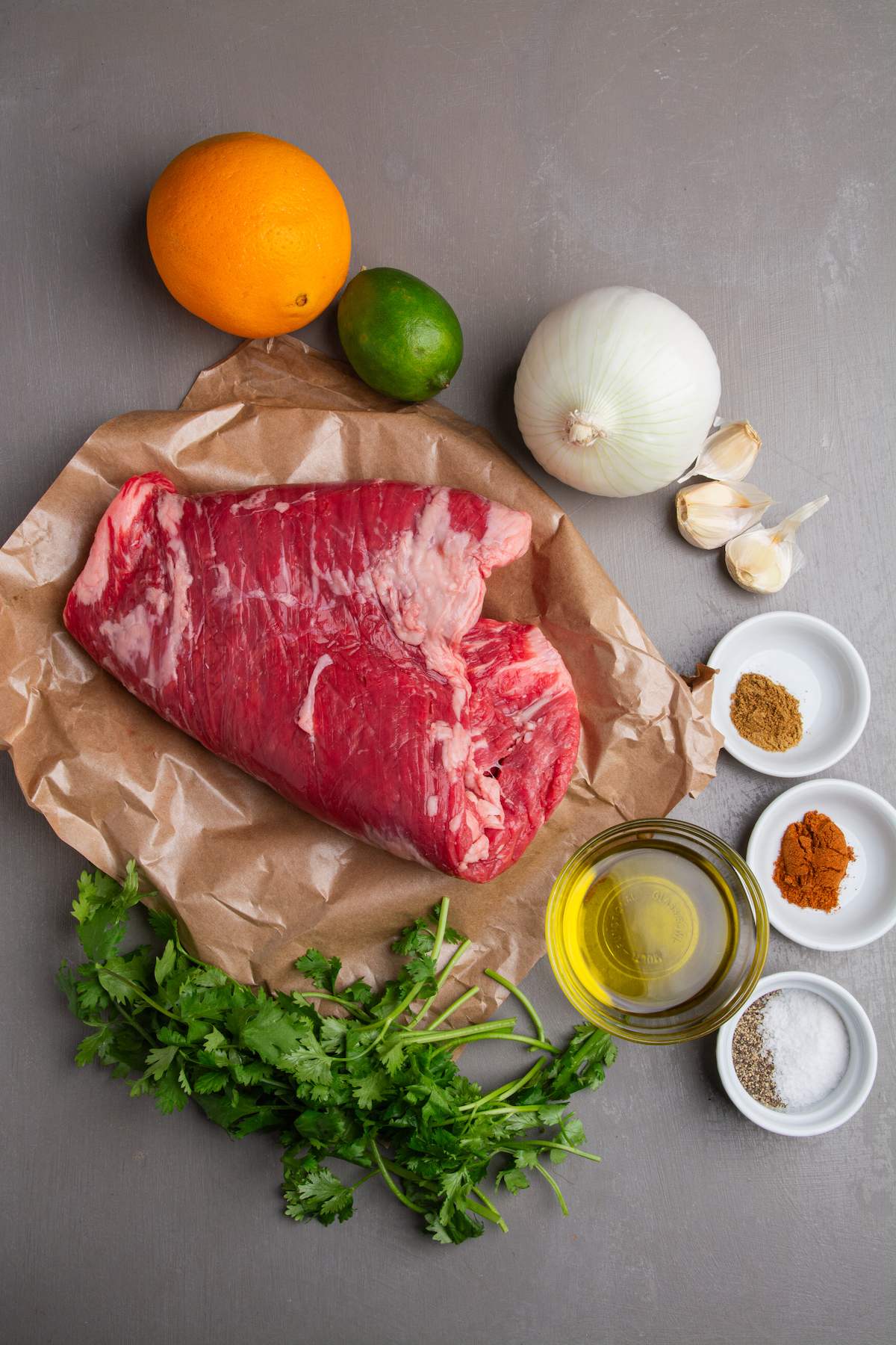 Ingredients for carne asada including flank steak, orange, lime, onion, garlic, cumin, chili powder, salt, pepper, oil and cilantro.