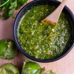 A bowl of roasted tomatillo salsa verde with a bunch of cilantro, half a lime and three tomatillos around it.