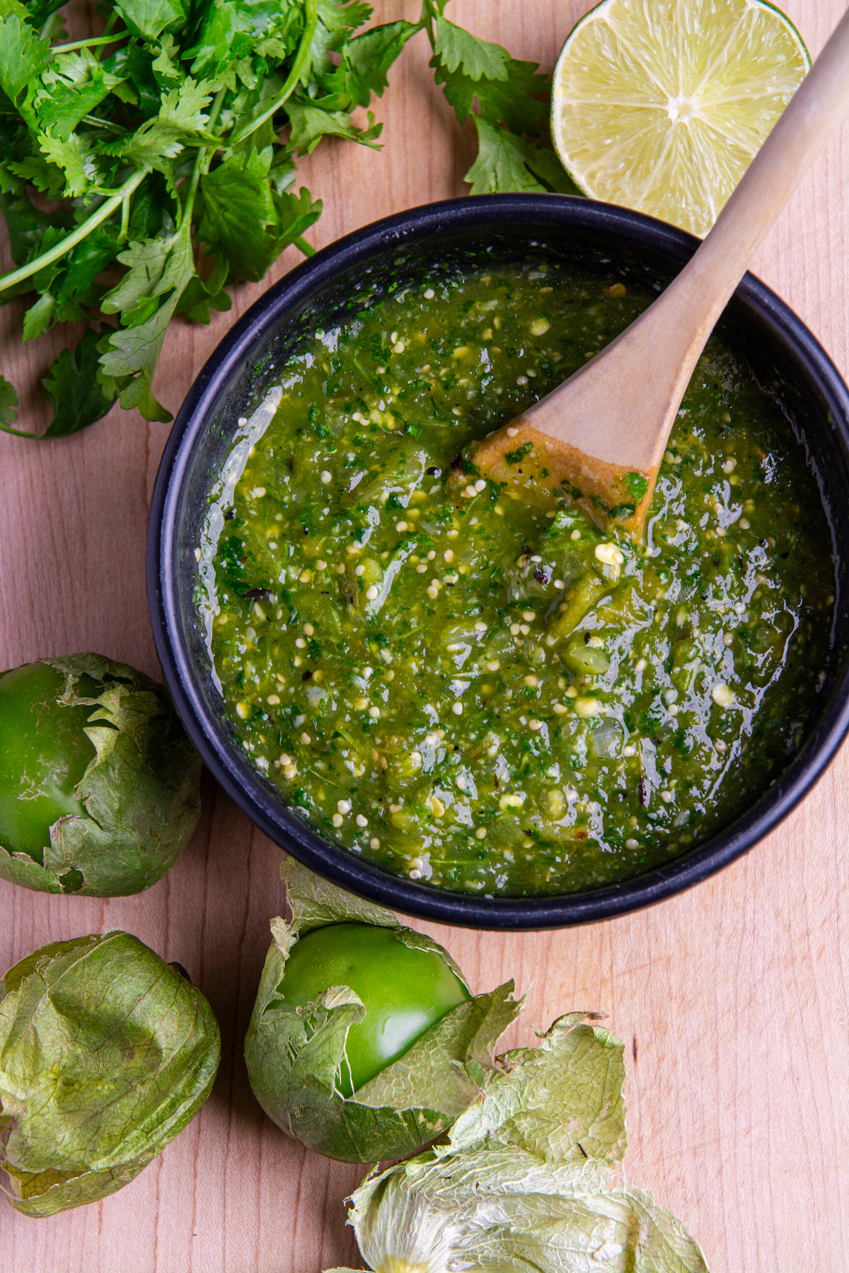 A bowl of roasted tomatillo salsa verde with a bunch of cilantro, half a lime and three tomatillos around it.