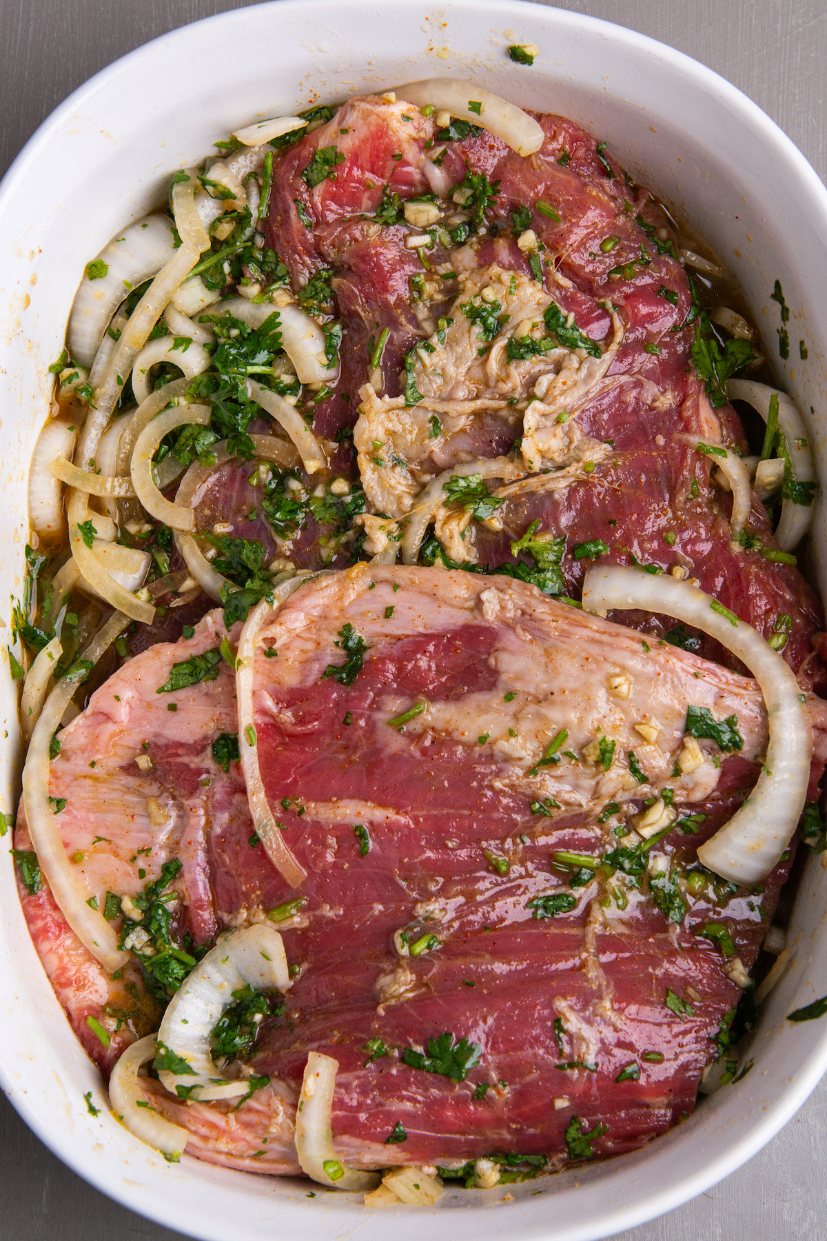 A deep ceramic roasting dish with two pieces of flank steak marinating in a carne asada marinade made with sliced white onion, chopped cilantro, and garlic.