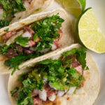 A plate with three carne asada tacos topped with cilantro, onion, and roasted tomatillo salsa verde, and two lime wedges placed on the right side of the plate.