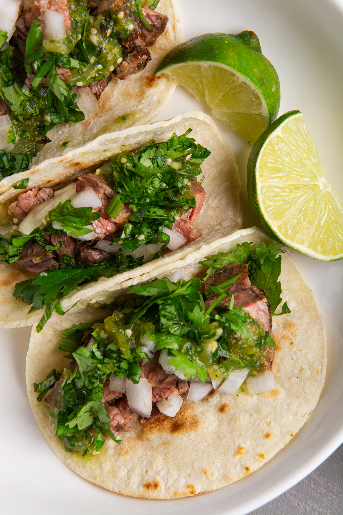 A plate with three carne asada tacos topped with cilantro, onion, and roasted tomatillo salsa verde, and two lime wedges placed on the right side of the plate.