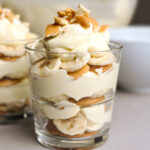 A glass of banana pudding with three layers of Nilla wafers, sliced bananas, vanilla pudding, topped with crumbles of Nilla wafers.
