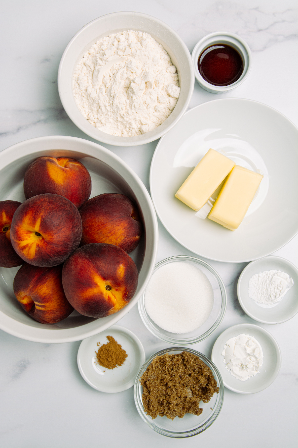 Ingredients needed for a peach cobbler: A bowl of fresh peaches, all purpose flour, a stick of butter, white sugar, brown sugar, ground cinnamon, baking powder, salt, and salt.