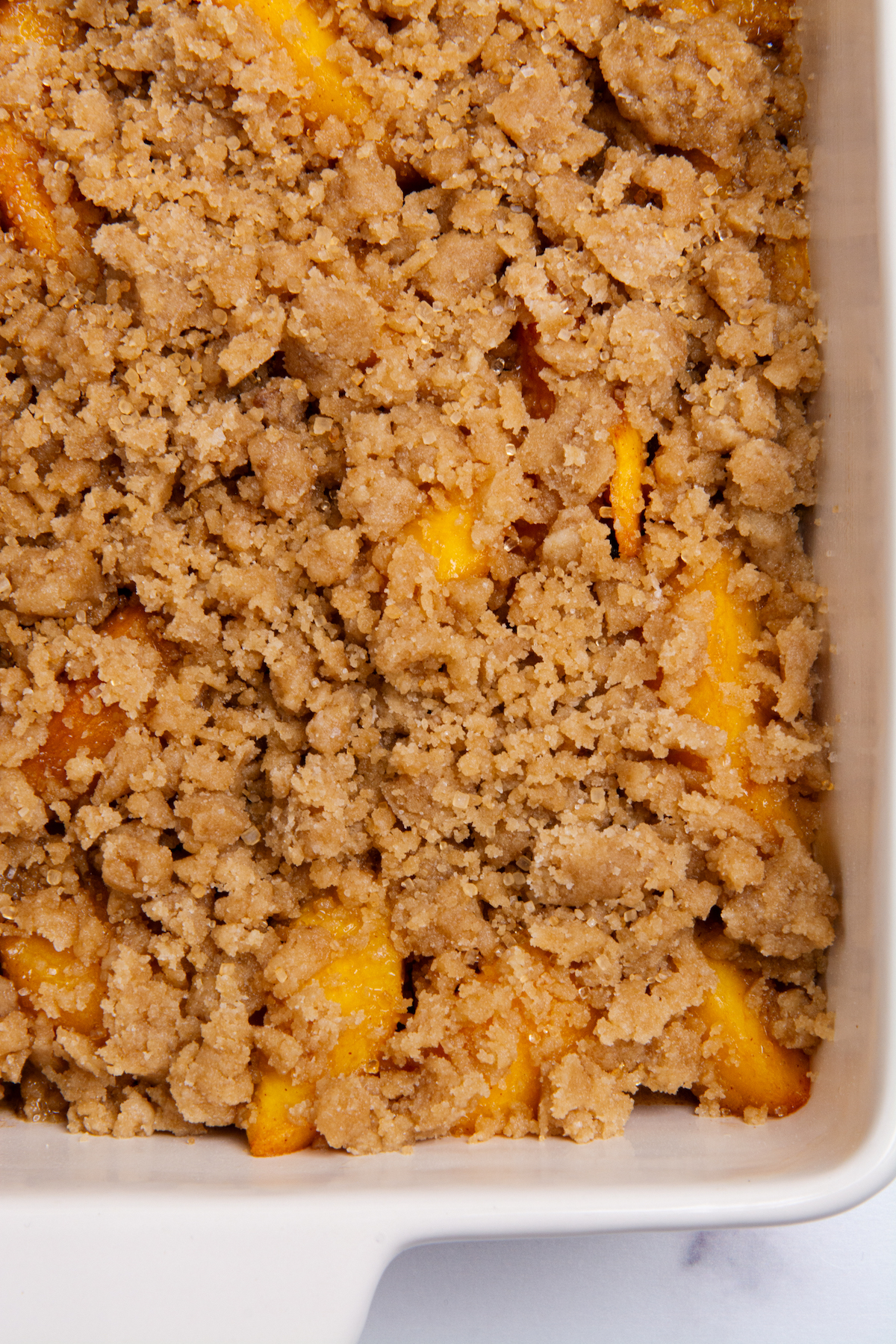 A baking dish with peaches coated with peach cobbler filling and topped with unbaked flour topping.