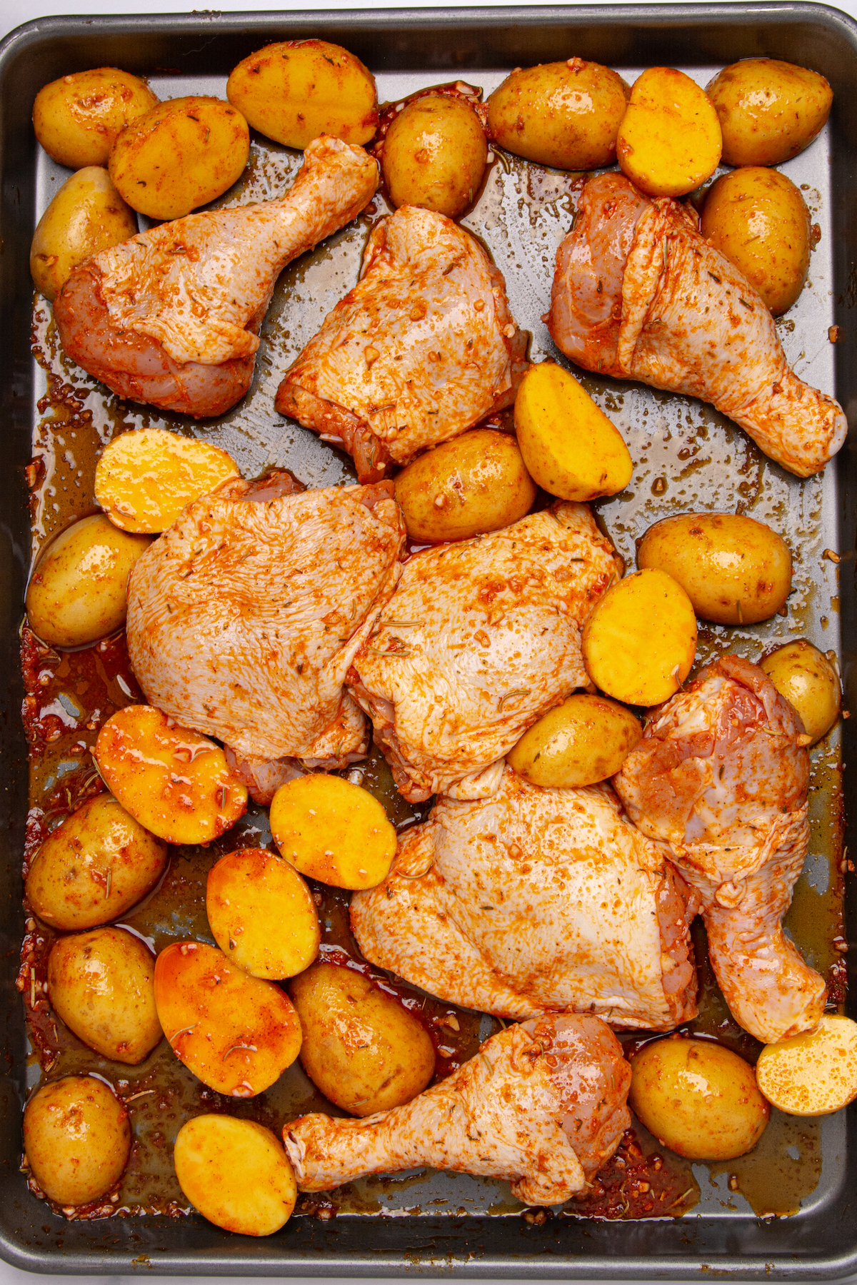 A baking sheet with raw chicken drumsticks, chicken thighs, and baby potatoes covered in sauce.