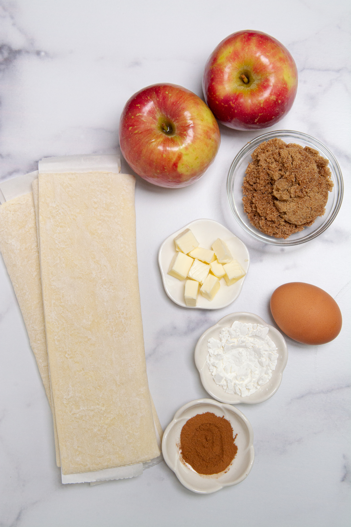 Ingredients for making apple turnovers, Fuji apples, frozen puff pastry, brown sugar, butter, cornstarch, salt, cinnamon, and egg.