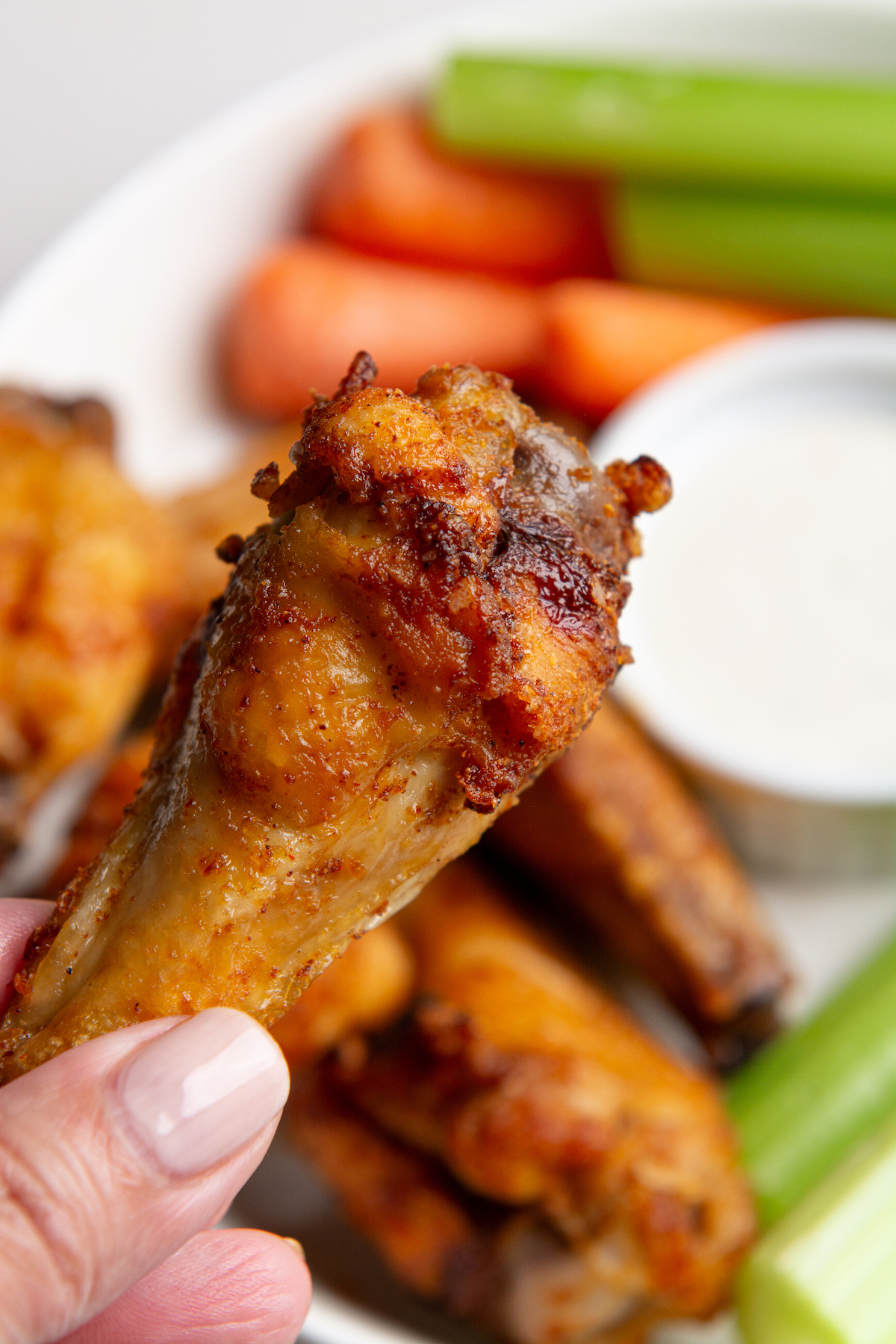 A hand holding a crispy, seasoned chicken wing drum with a plate of crispy chicken wings in the background.