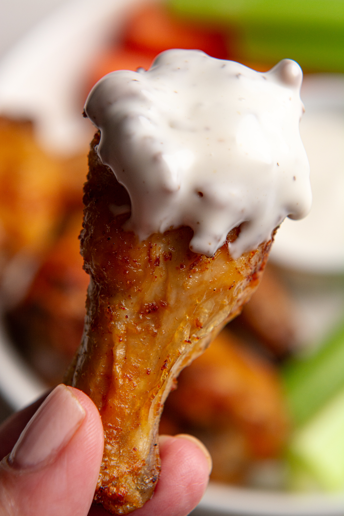 A hand holding a crispy chicken wing drum smothered in ranch dressing with a plate of crispy chicken wings in the background.