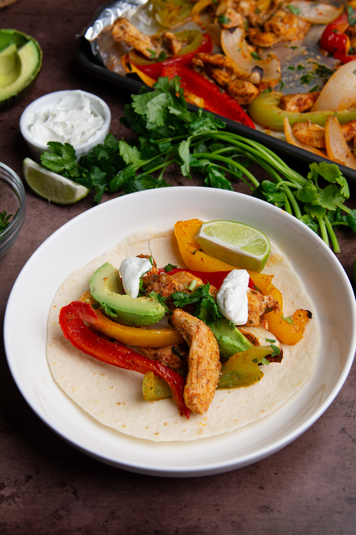 A plate with chicken fajitas served on a flour tortilla with a bunch of cilantro and a baking sheet of chicken fajitas in the background.