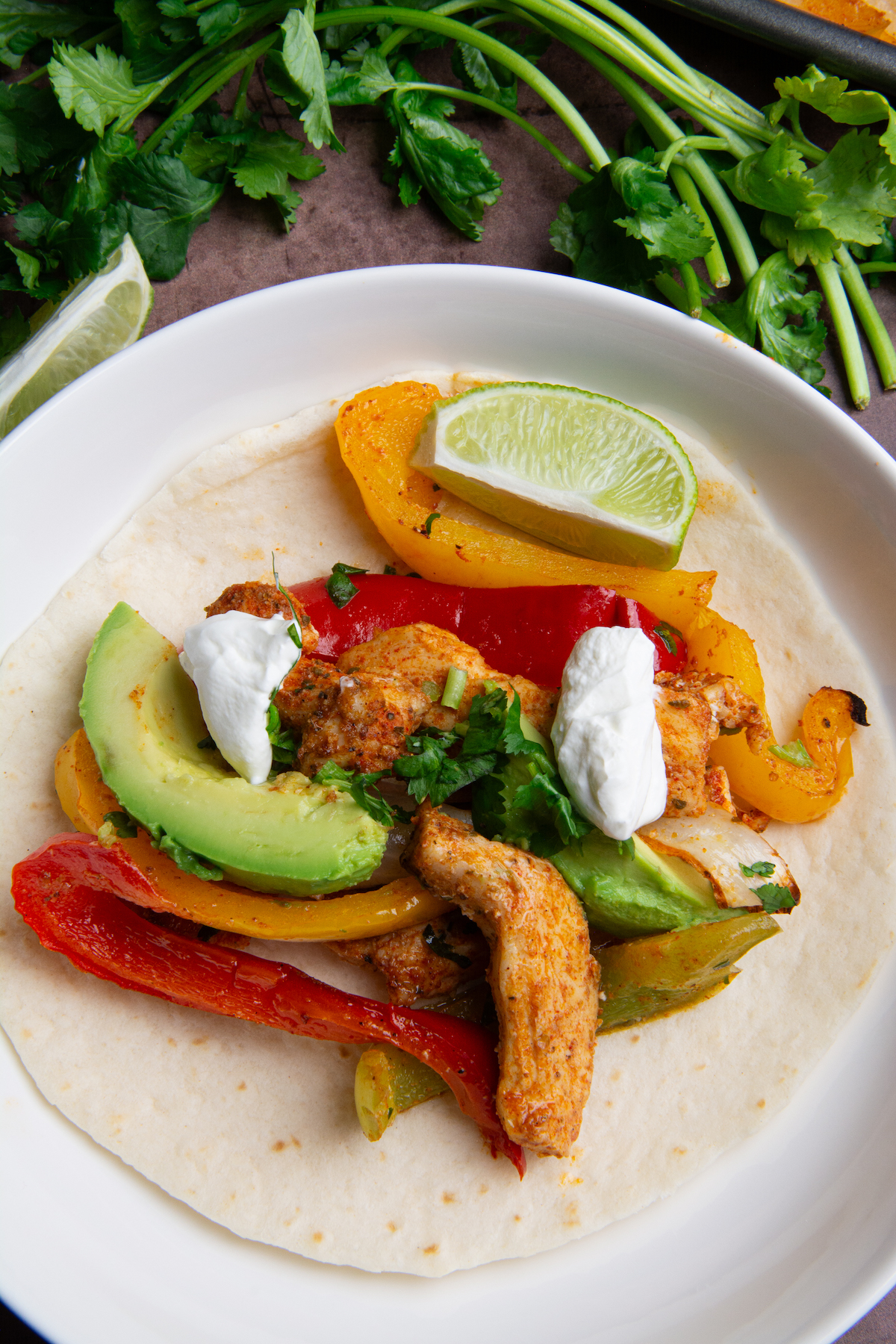 A plate with chicken fajitas served on a flour tortilla with a bunch of cilantro in the background.