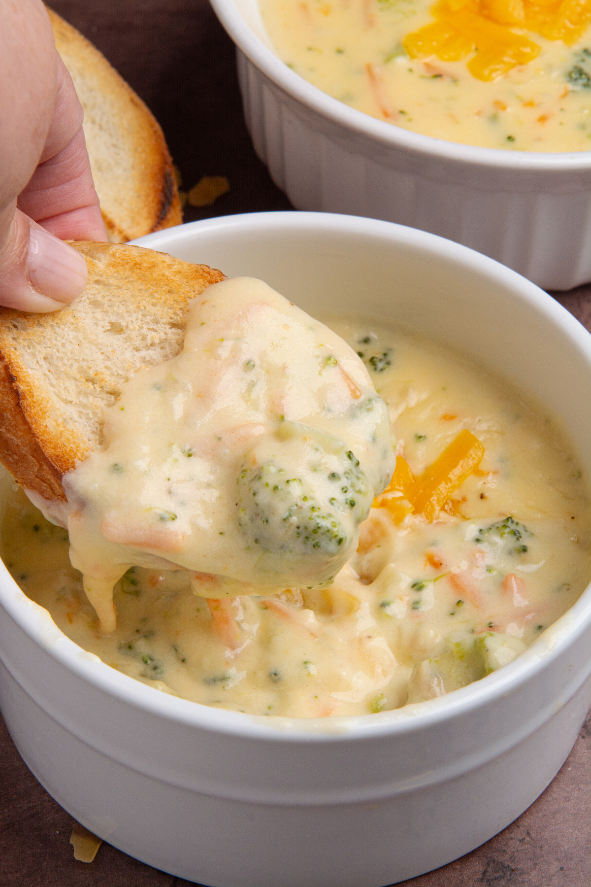 A hand holding a piece of toasted bread scooping out from a bowl of broccoli cheddar soup.