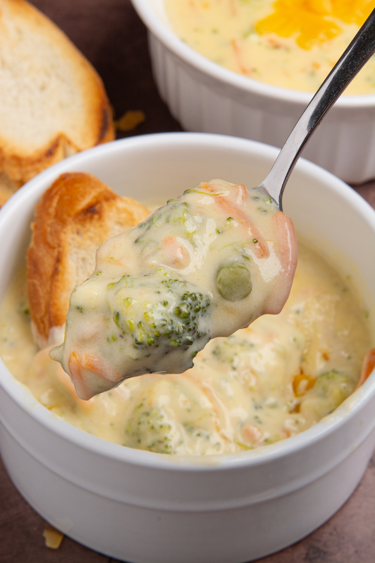 A spoonful of broccoli cheddar soup with a bowl of soup and a piece of toast in the background.