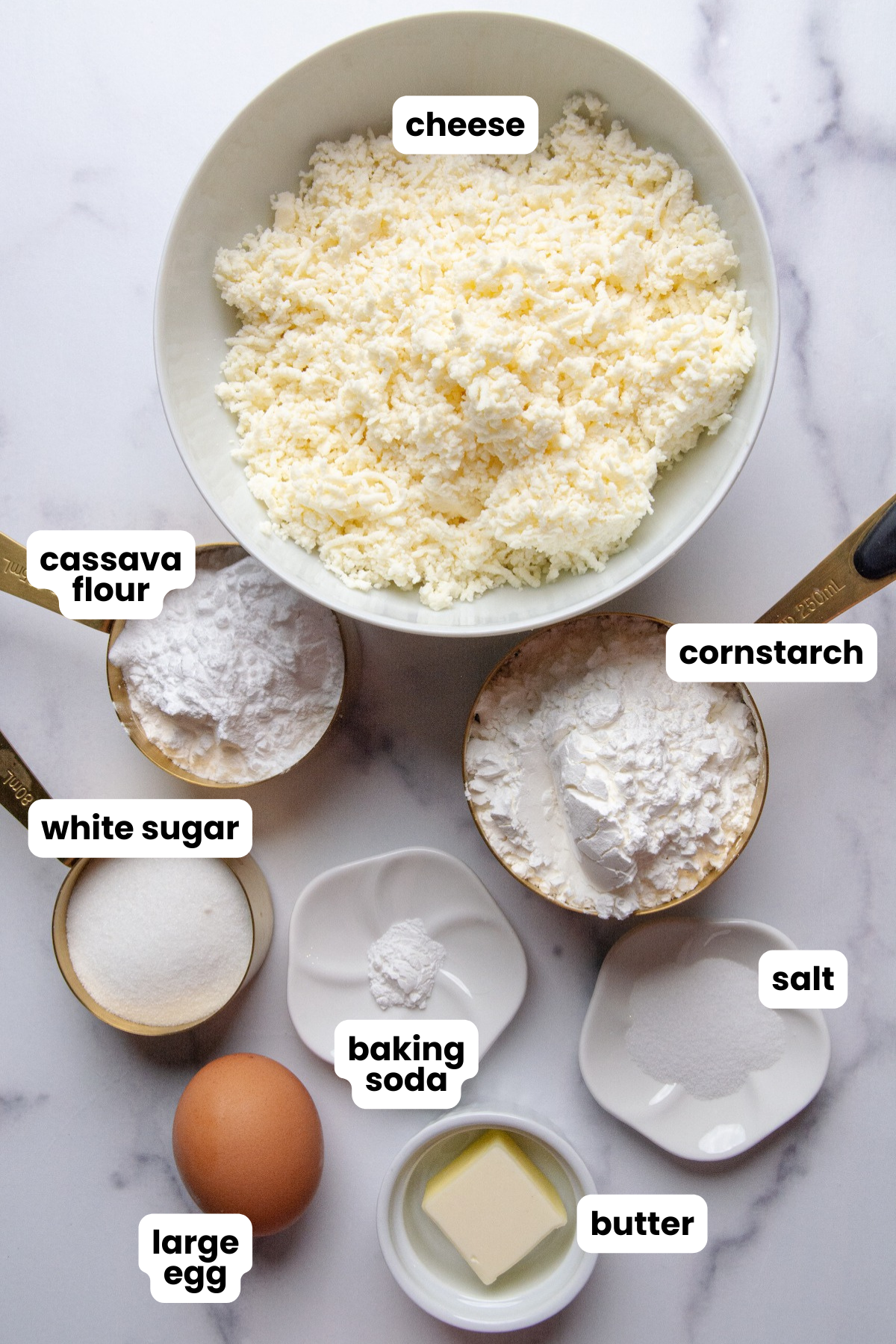 Ingredients for making Colombian buñuelos, cheese, cassava flour, cornstarch, white sugar, baking soda, salt, egg, and butter.