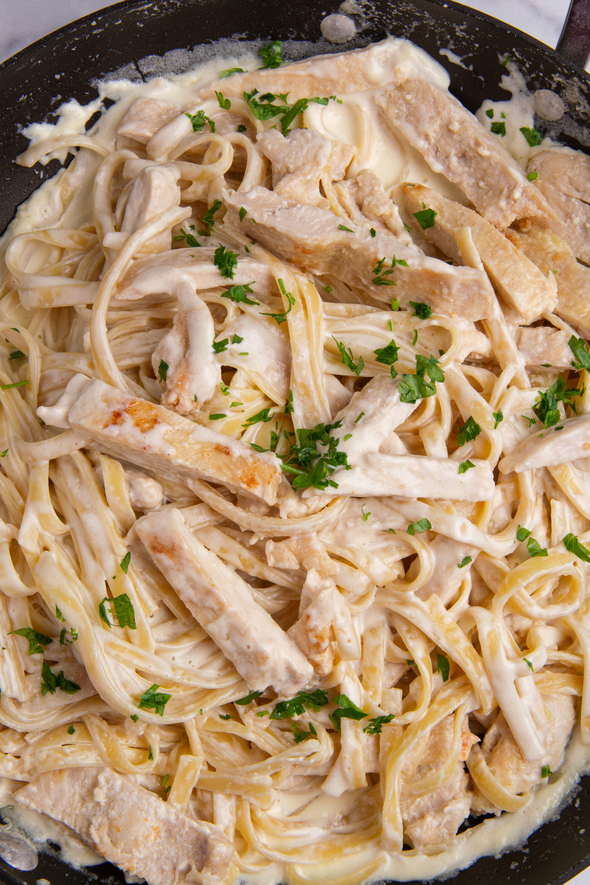 A large skillet with cooked Chicken Alfredo with fettuccine and a sprinkle of fresh chopped parsley.