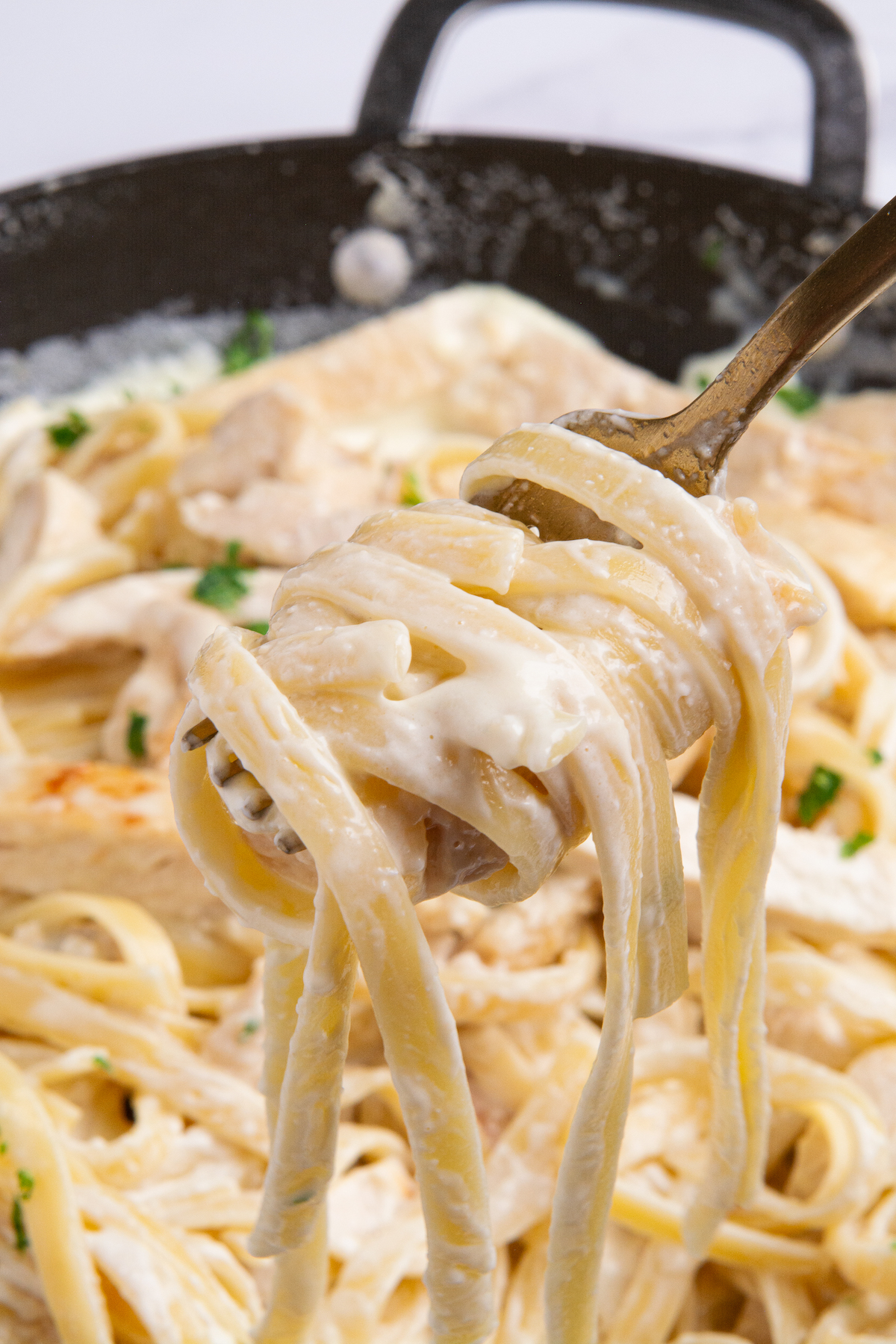 Fettuccine covered in Alfredo sauce twirled around a fork with a pan of fettuccine Alfredo in the background.