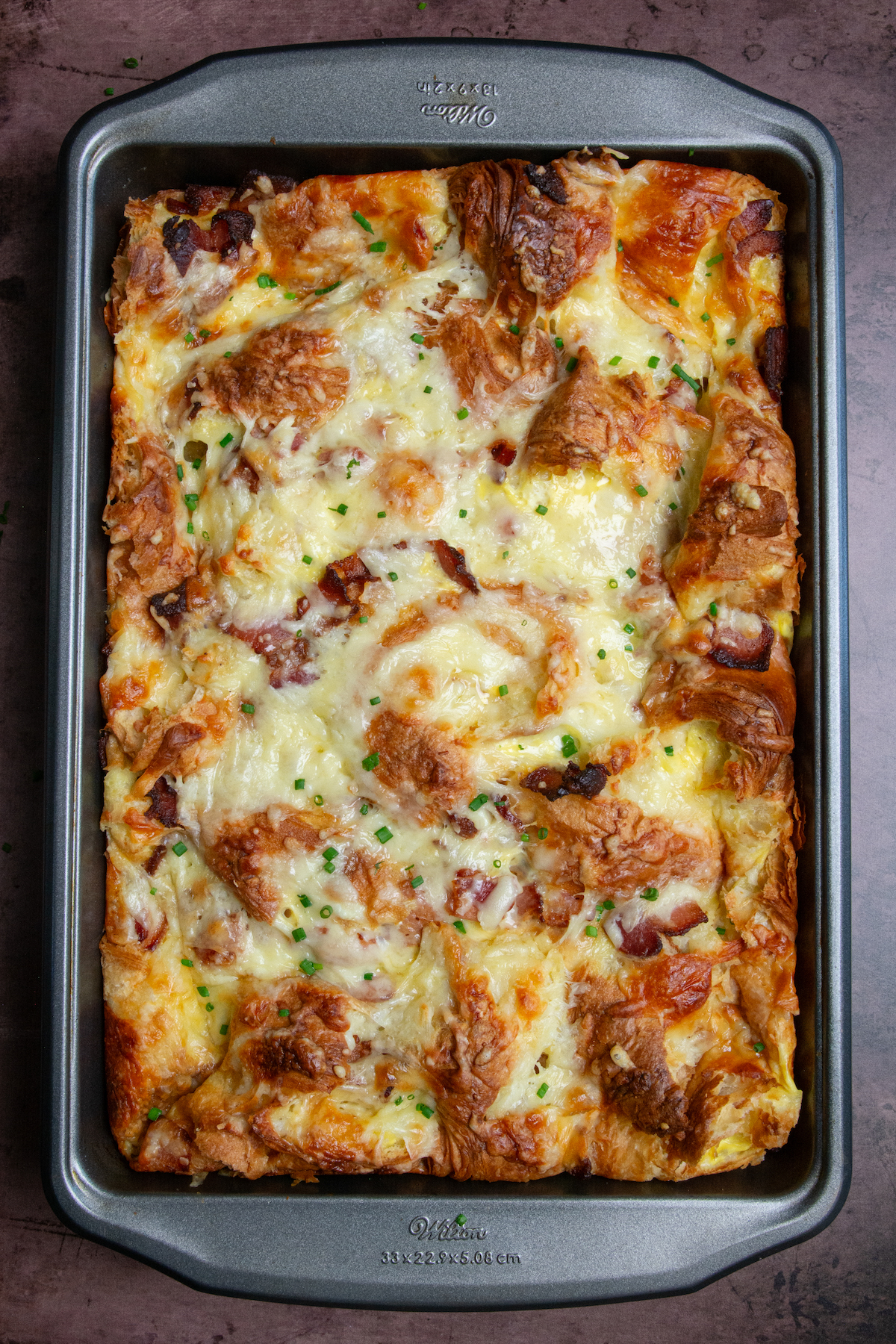 A golden brown croissant breakfast casserole with crispy bacon, melted cheese and garnished with chopped chives.