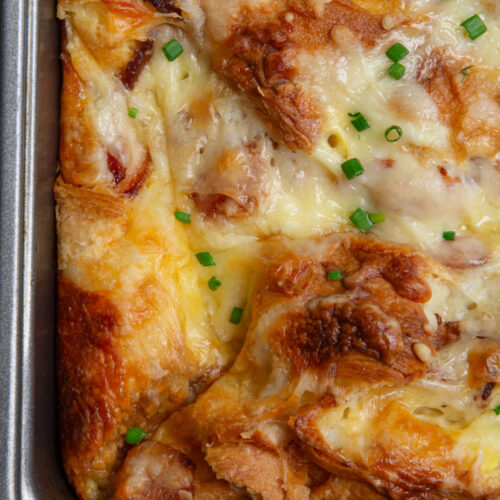 A corner of a golden brown croissant breakfast casserole with crispy bacon, melted cheese and garnished with chopped chives.