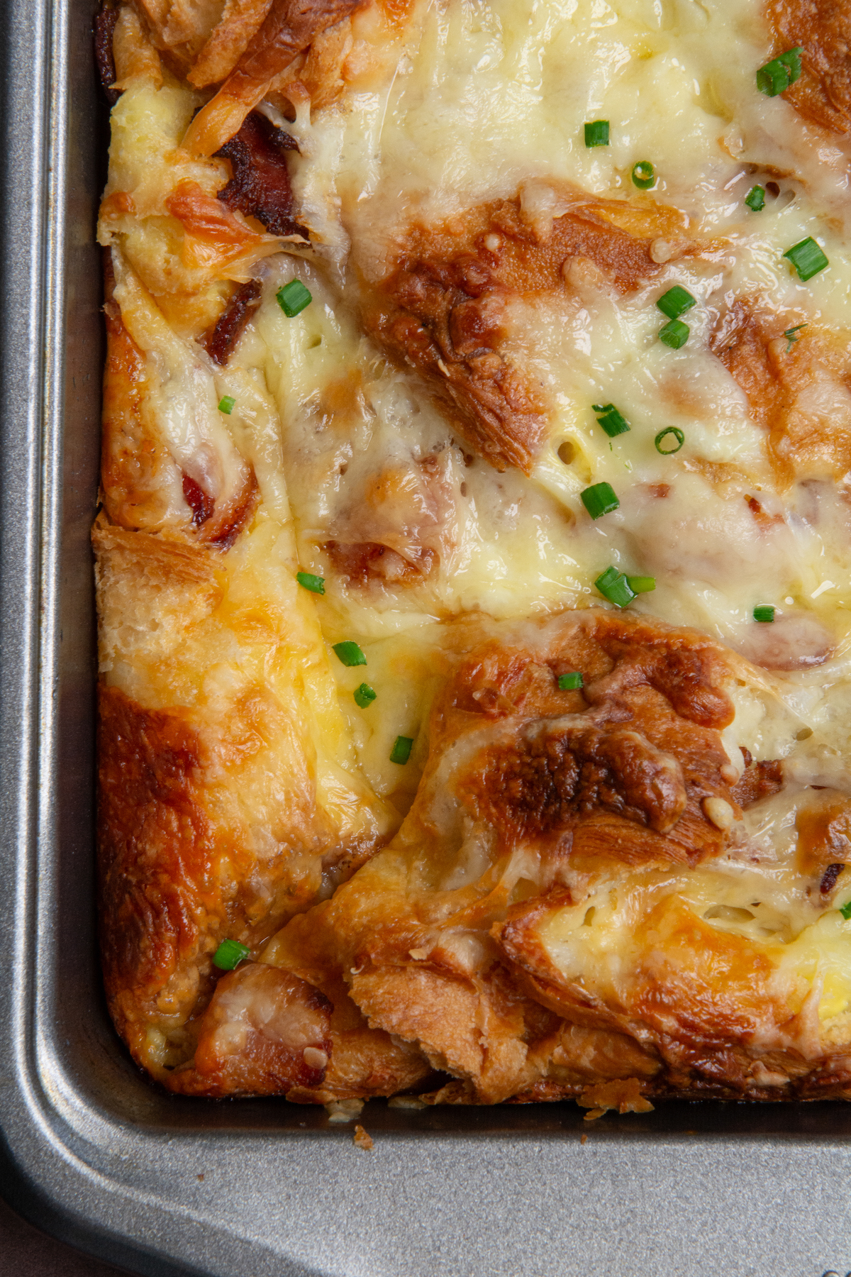 A corner of a golden brown croissant breakfast casserole with crispy bacon, melted cheese and garnished with chopped chives.