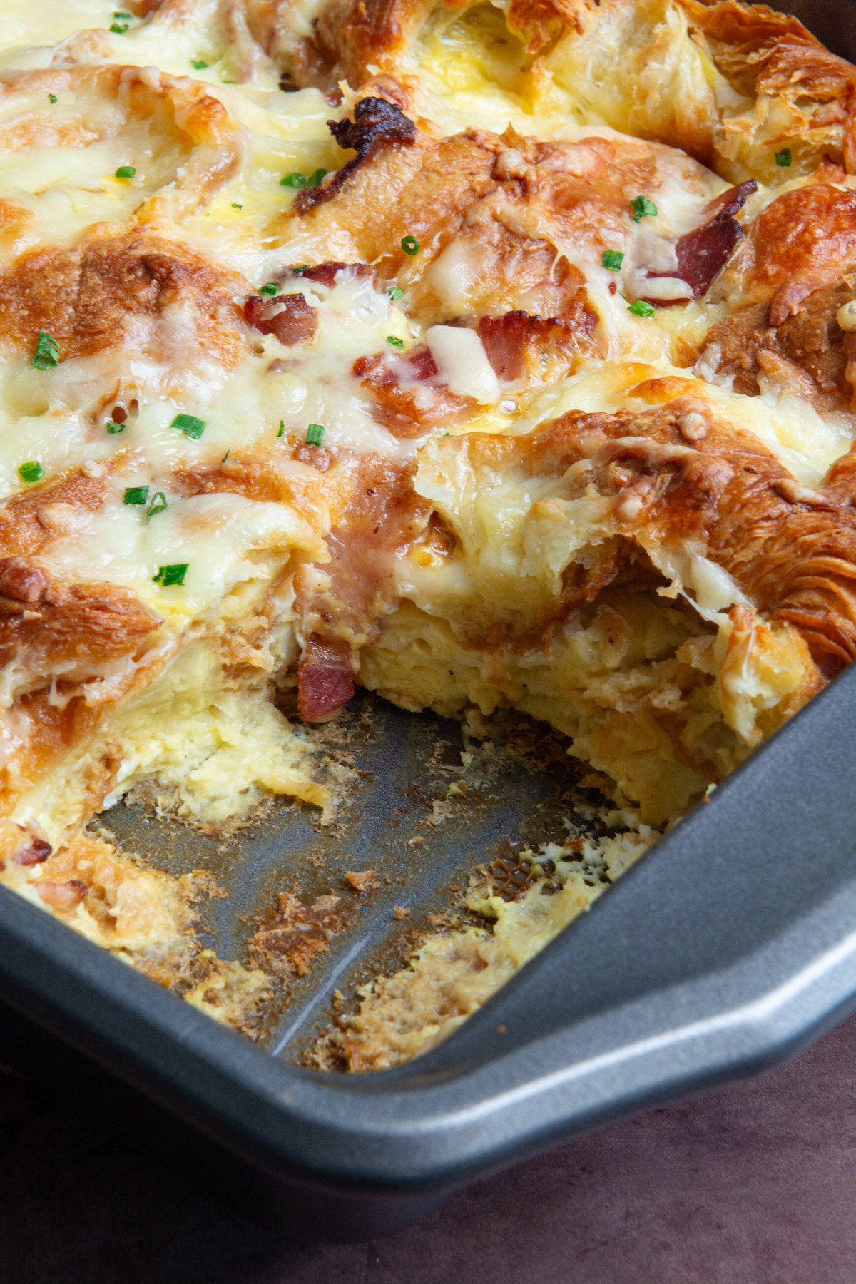 A corner golden brown croissant breakfast casserole with crispy bacon, melted cheese and garnished with chopped chives.