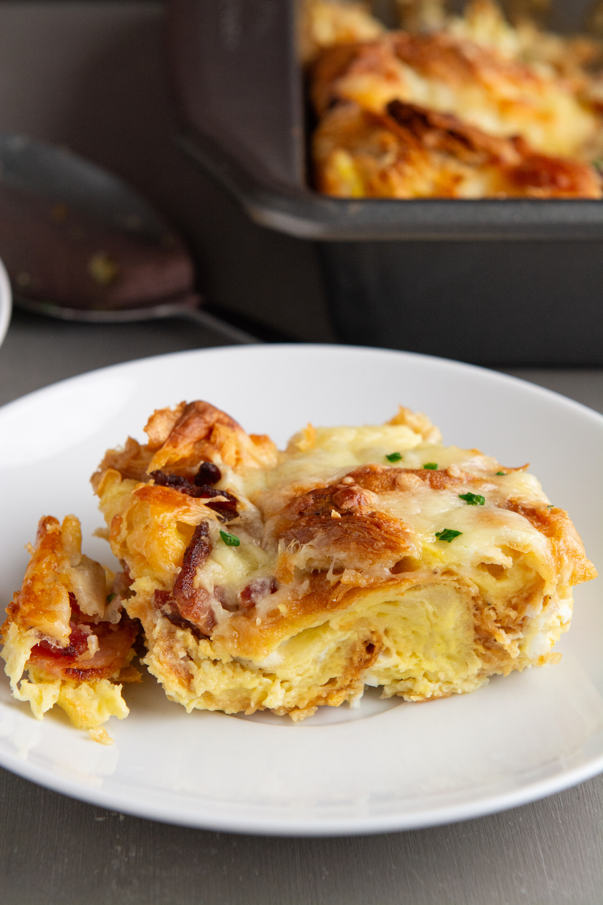 A slice of golden brown croissant breakfast casserole with crispy bacon, melted cheese and garnished with chopped chives.