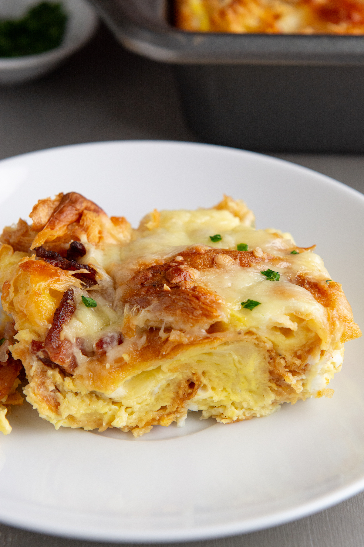 A slice of golden brown croissant breakfast casserole with crispy bacon, melted cheese and garnished with chopped chives.