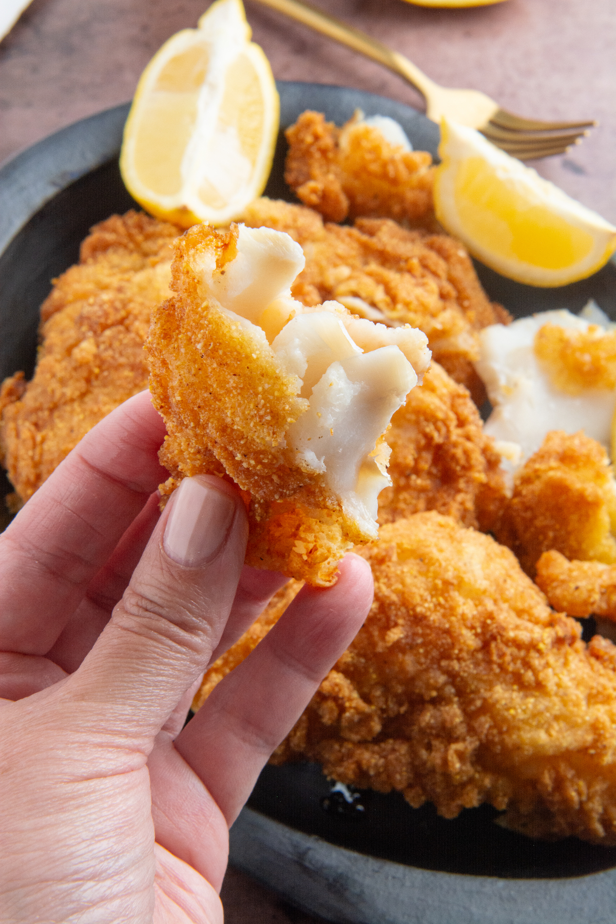 A hand holding a piece of crispy fried fish with a plate full of pieces of crispy fried fish and lemon wedges in the background.