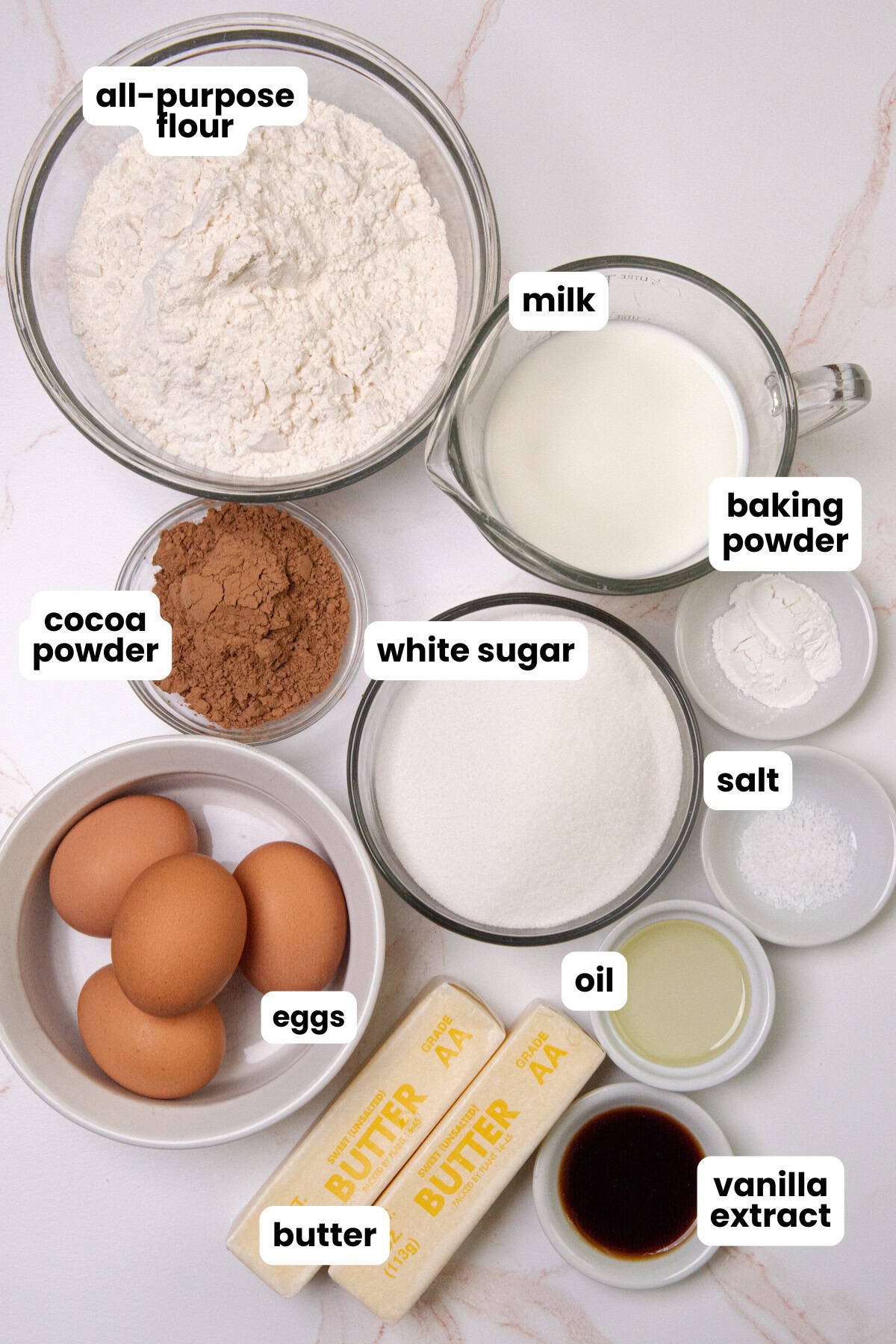 Ingredients for a marble cake recipe, all-purpose flour, cocoa powder, baking powder, white sugar, salt, butter, eggs, milk, oil, and vanilla extract.