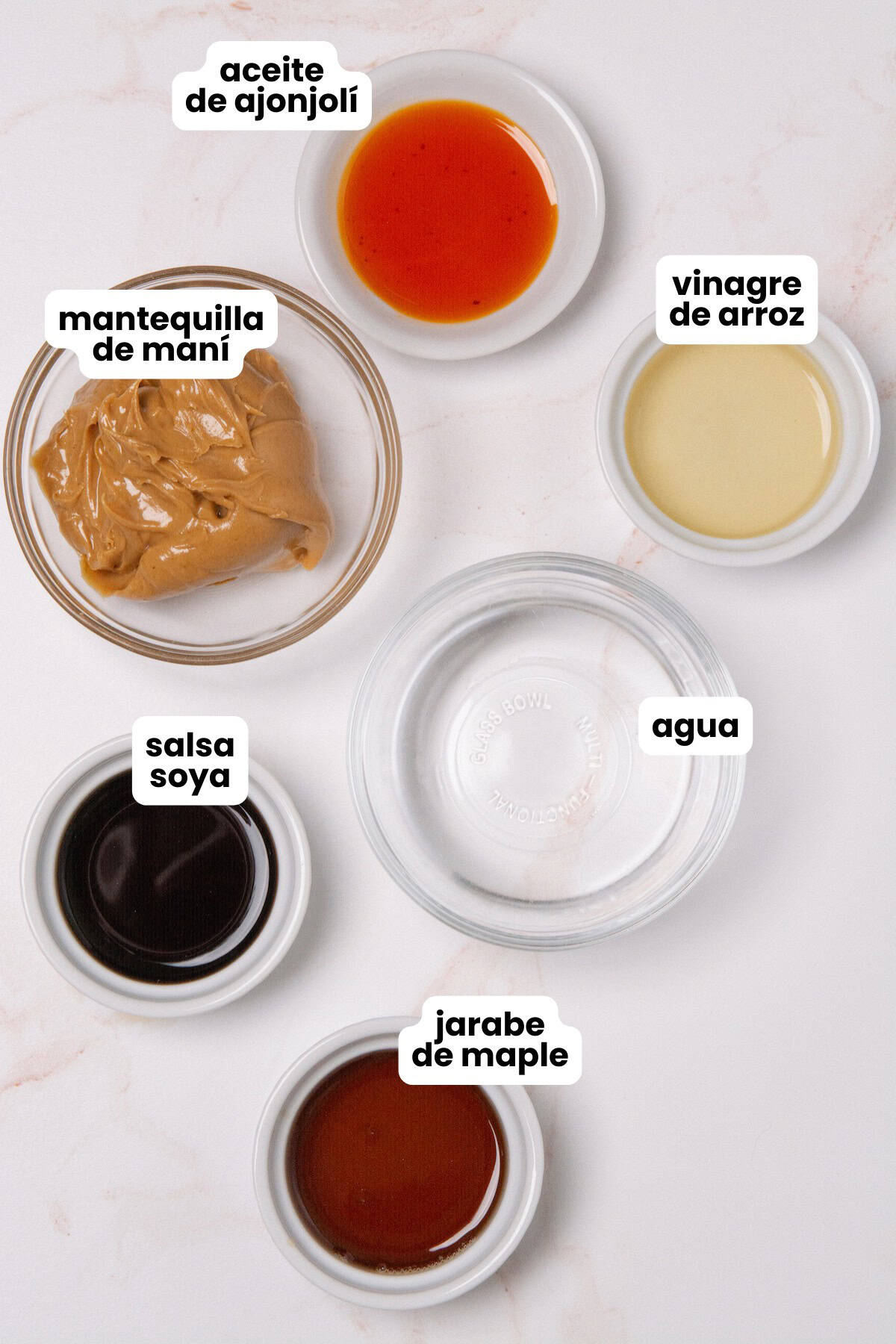 Ingredients in Spanish for making a creamy peanut sauce which include creamy peanut butter, rice vinegar, soy sauce, sesame oil, maple syrup, and water.