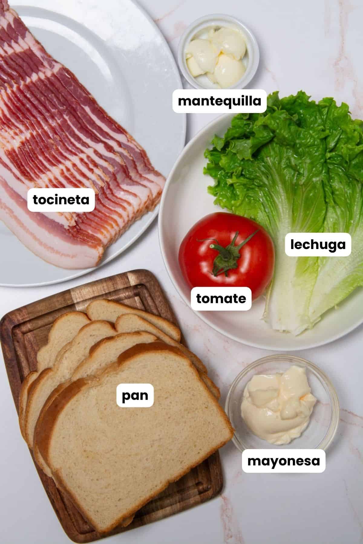 Inrgedients for a BLT sandwich in Spanish, bacon, lettuce, tomato, bread, butter, and mayonnaise.