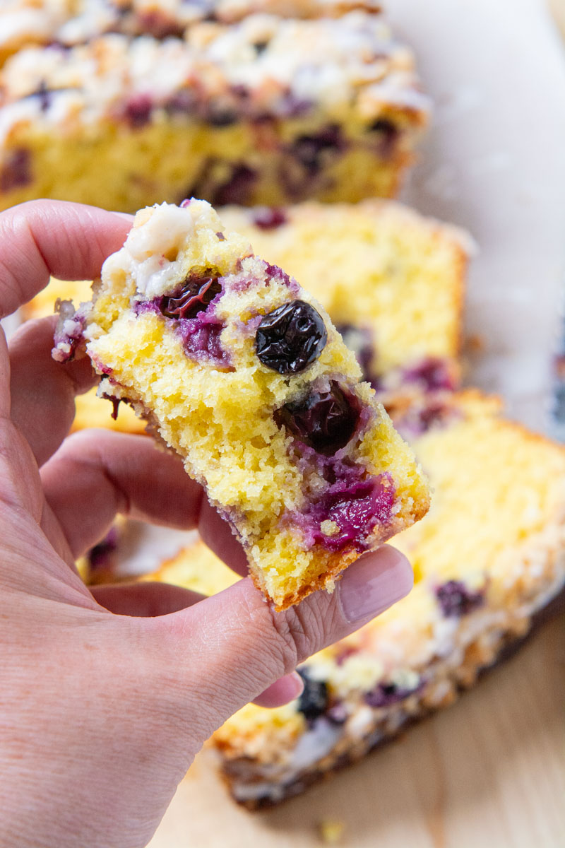 A hand holding a slice of a lemon blueberry loaf topped with a delicious, golden brown crumble and drizzled with a sweet lemon icing.