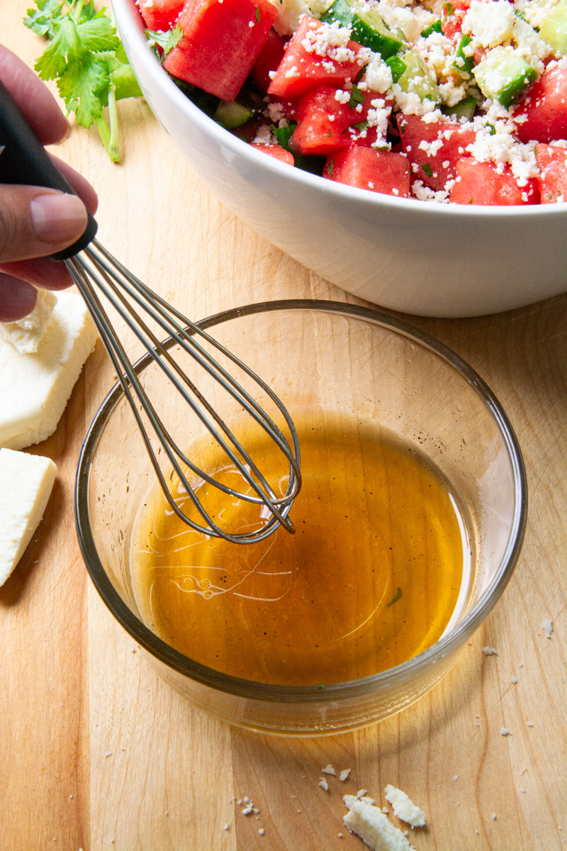 A small whisk mixing a sweet dressing for a watermelon cucumber salad made with maple syrup, lime juice, olive oil, salt, and pepper